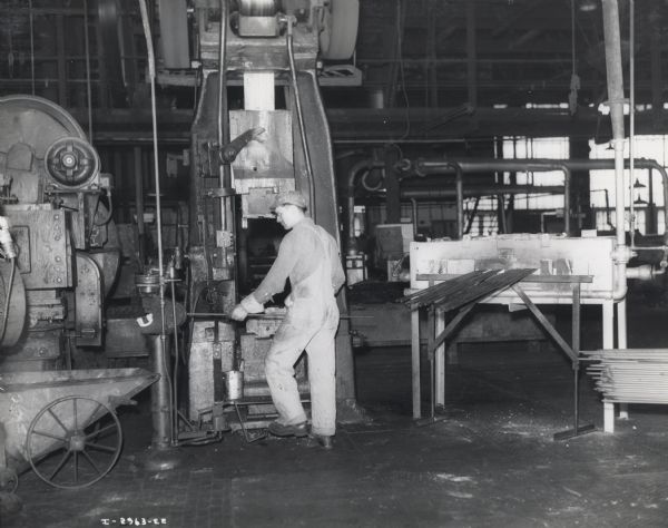 A factory worker operates a forge hammer at International Harvester's Canton Works. Original caption reads: "Shell Package Case Stops, Canton Works. Forge hammer and cut-off shear operated in manufacture of shell stop packings. The operator is cutting and forging off the end of the bar, which he holds in his hands. Note the hot forging falling into the truck at the rear of the shear."