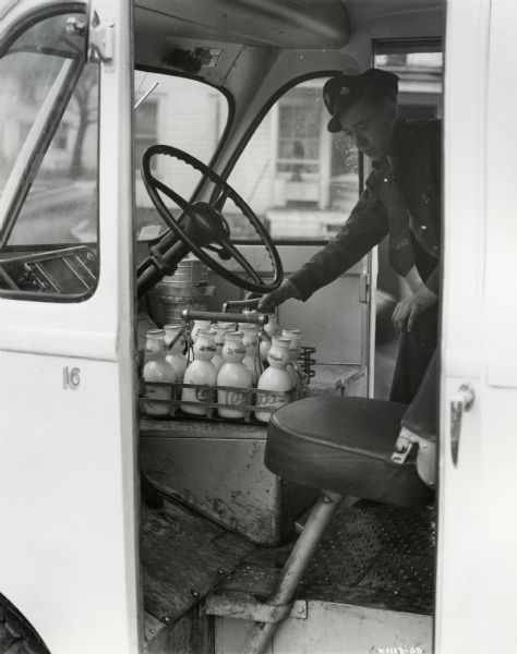 Cab of an International model D-15-M truck with 102 inch wheelbase and Metro body. The delivery man is standing inside the cab on the passenger side of the van, removing a basket of milk bottles.