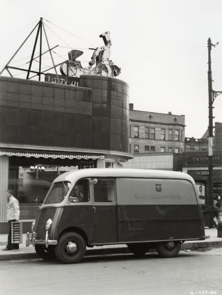 Man standing with a pie container next to an International model D-15-M truck with Metro body. According to the original caption, "Chef Wright" is the driver and the truck could carry 444 crust pies and 193 cream pies. The cream pies were stored in insulated and refrigerated compartments. On top of the nearby building is an electric sign of a chef riding a horse, which is pulling a carriage.