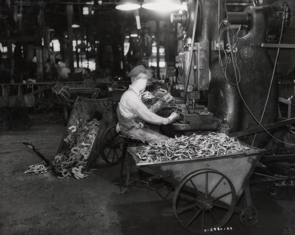 A factory worker trims forgings at International Harvester's Canton Works. Original caption reads: "Shell Package Case Stops, Canton Works. This operation shows the flash trimming of package forgings on a trimming press. The truck at the left of the operator shows the forgings before the trimming operation and the truck at his right shows the forgings after the trimming operation is performed."