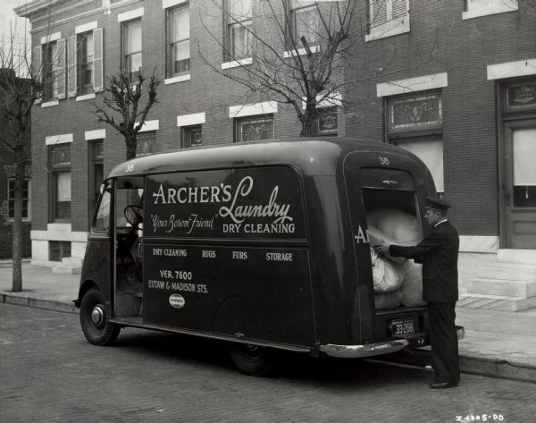 A man in a uniform standing at the back of an International Metro truck owned by Archer's Laundry [and] Dry Cleaning. The side of the truck includes the slogan: "Your Bosom Friend." A building with stained glass windows is in the background.
