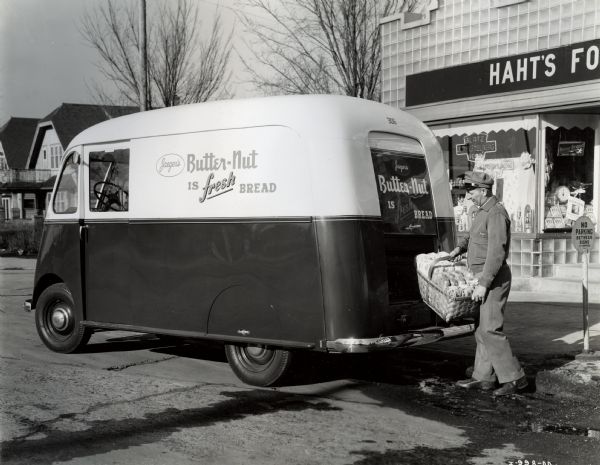A man standing at the back of an International Metro truck with a basket of bread. The truck includes the text: "Jaeger's / Butter-Nut is fresh bread". "Haht's" food(?) store is behind on the right and houses are on the left.  Near the man is a "No Parking" sign.