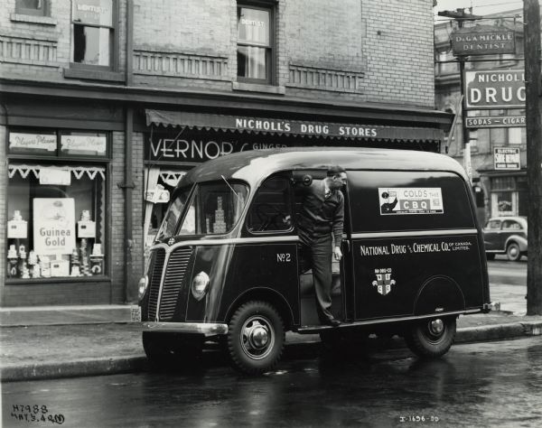 A man standing near an International Metro truck owned by the National Drug and Chemical Company. Nicholl's Drug Store is in the background, with a sign for a dentist in the window on the second floor.