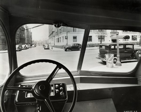 View from the driver's seat of an International Metro truck, showing part of the steering wheel and a city street beyond. Automobiles are parked near the corners, and there appears to be a park at the end of the street. A street sign on the right reads: "West End Ave".