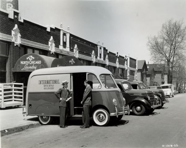 Mr. Kuss of International Harvester's Chicago North Side Branch with an International model D-2-M truck with a 102-inch wheelbase and a Metro body. Mr. Kuss is showing the truck to one of the owners of the North Chicago Laundry, which is in the background. Several houses can be seen further down the block. Cars and trucks are parked along the road.