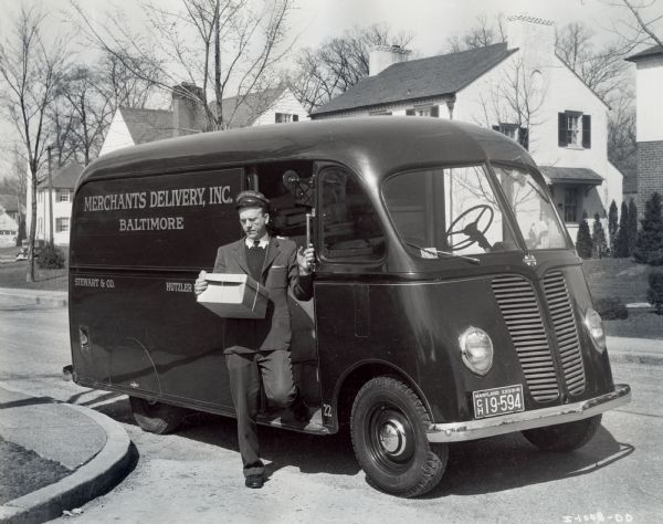 Uniformed driver stepping down from an International model D-2-M truck with a 113-inch wheelbase and a Metro body in a residential neighborhood. According to the original caption, Merchants Delivery owned two Metros and eight International trucks in all. The company delivered packages for two of Baltimore's leading department stores: Hutzler Brothers, and Stewart & Company.