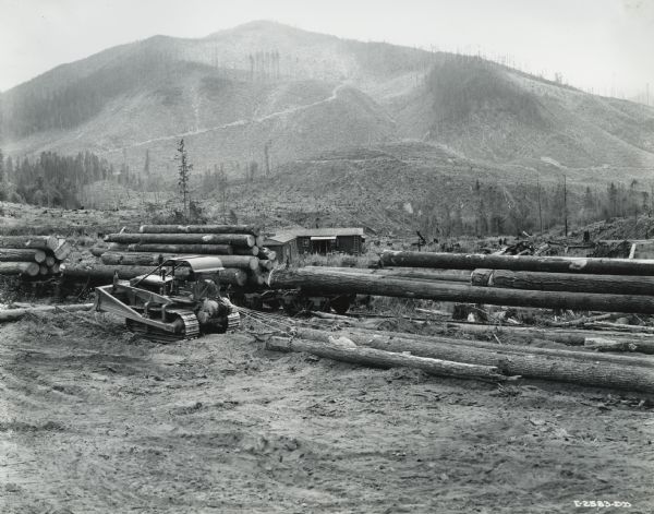 A man pulling logs with an International TD-18 Diesel TracTracTor (crawler tractor). The tractor was owned by the Pacific National Lumber Company and was engaged in selective logging of red fir on its property seven miles from its mill in National, Washington. Foothills and a mountain in the background show evidence of deforestation.