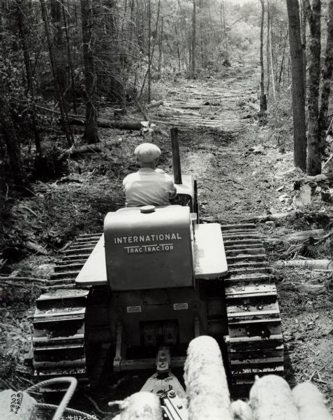 Rear view of a man pulling logs with an International TracTracTor (crawler tractor) on a logging road in a forest.