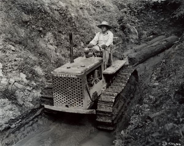Seldon Starcher, owner and operator of an International TD-6 Diesel TracTracTor (crawler tractor), uses his tractor both in logging and oil field work. He wears a straw hat as he drags logs down a muddy path with his tractor.
