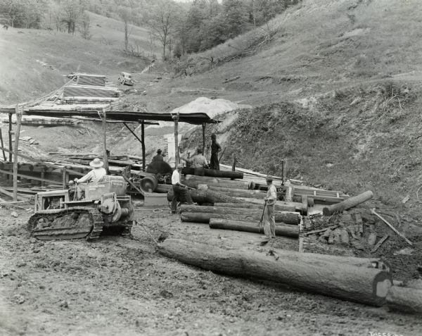 Seldon Starcher, owner and operator of an International TD-6 Diesel TracTracTor (crawler tractor), uses his tractor both in logging and oil field work. Starcher pulls logs behind the tractor to an area where men are working with a belt-driven saw. An automobile is parked on a hill in the background.