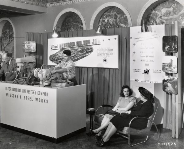 A blind(?) man operates a piece of machinery behind a table in a display booth at the "blind veterans show." The booth is labeled "International Harvester's Company / Wisconsin Steel Works" and a sign behind the man reads: "Blind Veteran on the Job." Two women are sitting in chairs to the man's left.