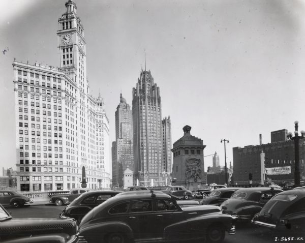 Numerous cars driving on what appears to be the Michigan Avenue Bridge (later renamed DuSable Bridge) in front of the Wrigley and Tribune buildings. There is a clock tower on the Wrigley building, and a bas-relief sculpture on the bridge. In the background on the right, a banner is hanging from the side of a warehouse building for Hibbard, Spencer, Bartlett & Co. that reads, in part: "Join Us In Celebrating Harvester's 100 Years in Chicago / South End Soldier Field Oct 18 - Nov 2."