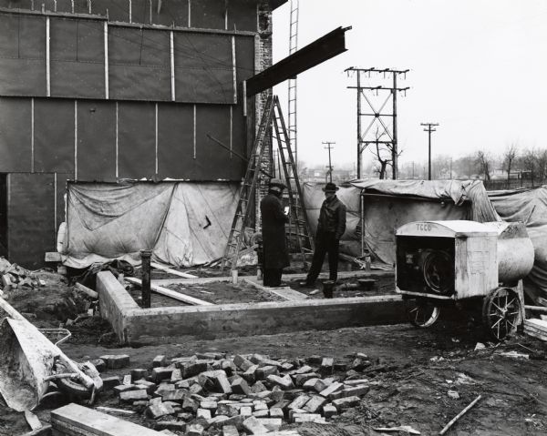Two men talking on a construction site, probably at International Harvester's Bettendorf Tank Arsenal (factory). A pile of bricks is in the foreground along with a wheelbarrow, ladder, cement-mixer and other building materials.