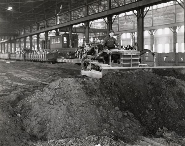 A man operates an International crawler tractor (TracTracTor) with a Bucyrus-Erie bulldozer blade inside a factory building. The building is probably International Harvester's Bettendorf Tank Arsenal.