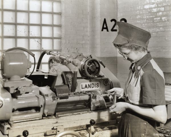 A female factory worker operates a grinding machine. Original caption reads: "This woman employee on torpedo production operates a grinding machine. The part she is shown measuring with a micrometer must be machined to a tolerance of .0005 of 1/2 thousandths of an inch. Trained in a special course for this work, she had only a few months' prior experience as a machine operator. She sets up her own machine for the work."