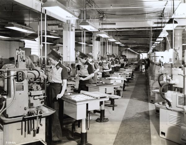 A line of female factory workers operate milling machines at an International Harvester plant responsible for manufacturing aircraft torpedo parts. Original caption reads: "A series of small milling machines, all operated by women employees, milling small parts for the torpedo."