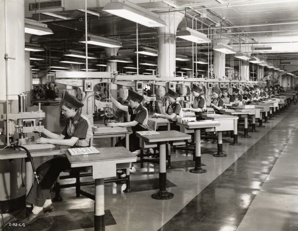 A long line of female factory workers operating drill presses in an International Harvester aircraft torpedo plant. Original caption reads: "A battery of 16 small, single spindle, sensitive drill presses operated by women employees. Women are especially adaptable at this type of machine operation because their sensitive fingers and delicate feel and touch enable them to handle the small parts more skillfully than men operators."