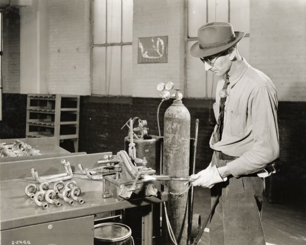 A factory worker uses an oxy-acetylene torch to fit a copper tube in the production of an aircraft torpedo. He is wearing an International Harvester Company / McCormick button on his shirt. Original caption reads: "A great amount of copper tubing is used in the construction of the torpedo, much of which must be bent and fitted to precise dimensions. In order to do this work by mass production methods, the company adopted the use of an acetylene heat flame. The operator holds the flame on the tubing and slowly bends it to fit a master jig, as shown in the photograph. This method has greatly speeded [sic] up the operation."