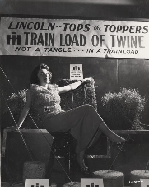 Marilyn Schultz of International Harvester's Lincoln office models a sweater made of twine as part of a district office promotional event. A sign above reads: "Lincoln Tops the Toppers, IH Train Load of Twine, Not a Tangle in a Trainload." Original caption reads: "Adding female pulchritude to the Lincoln, Nebraska, district office twine exhibit in a home-made International Harvester twine sweater is Marilyn Schultz of the local office."