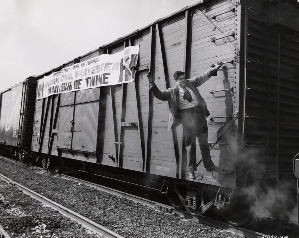 A man hangs on to the side of a train from a ladder and waves his hand. Original caption reads: "Giving his engineer the 'highball' upon arrival of the 25-car train load of twine in Lincoln, Nebraska, is Robert Sargent, International Harvester sales promotion supervisor at Lincoln." Banner on side of trains says: "Lincoln District Tops the Toppers / International Harvester / Railroad of Twine."