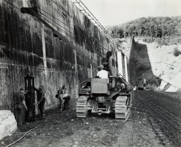 Men work with an International TracTracTor (crawler tractor) in construction of the Merriman Dam. The tractor was owned by S.A. Healy Co., White Plains, New York. Original caption reads: "IHC tractor with Superior Swing Crane (Trackson pipe-laying crane), swinging air-driven earth tamping hammer. Tamping earth fill next to concrete core on construction of Merriman Dam, in Catskill Mountains."