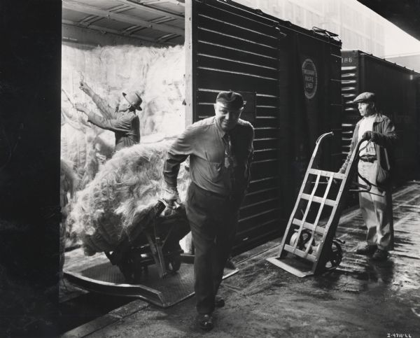 Three factory workers unload bundles of sisal fiber (fibre) from a train at an International Harvester twine mill. A note with the companion photo to this image reads: "Unloading fiber at an International Harvester twine mill from various parts of the world -- from Mexico, Cuba, Africa, Brazil, Haiti, the Philippine Islands, Central America, and elsewhere. This variety of sources provides fibers to produce the proper blends required to make baler and binder value to the IH quality standard."