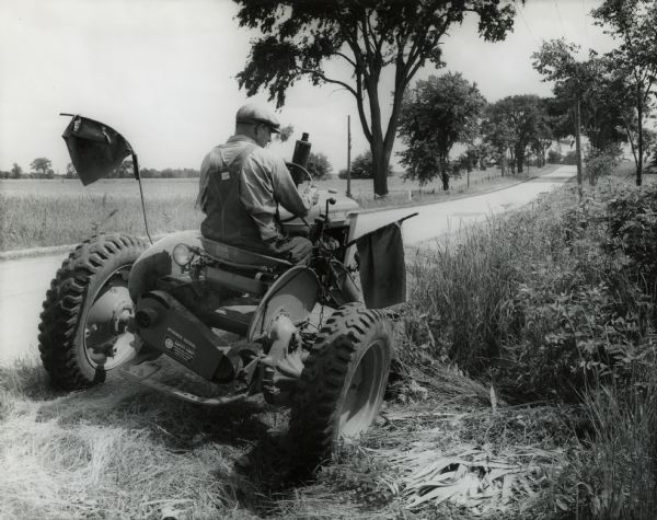 Man mows grass with an International A tractor and mower. Caution flags are mounted on the tractor. Original caption reads: "International A used with an International mower cutting heavy grass and weeds along the highway."