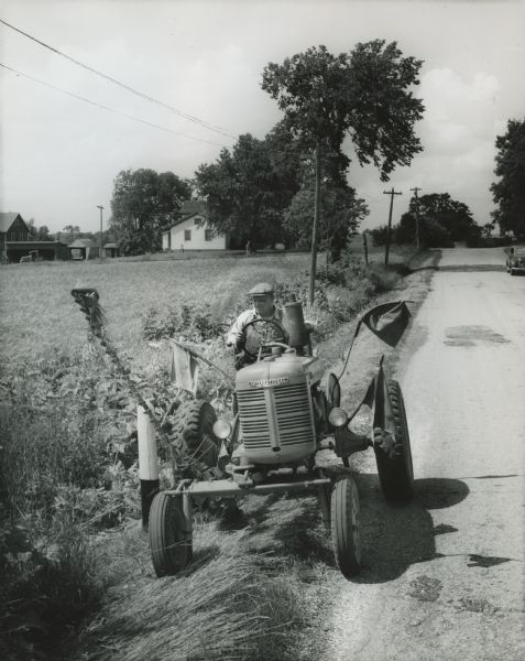 Man mowing grass along a road or highway with an International A tractor and mower. A house, barn, some sheds, a car, and a tractor are in the background, and a person is walking down a driveway. Caution flags are mounted on the tractor. Original caption reads: "The International A with International mower. The mower is being raised over a guard post. This operation required about three seconds."