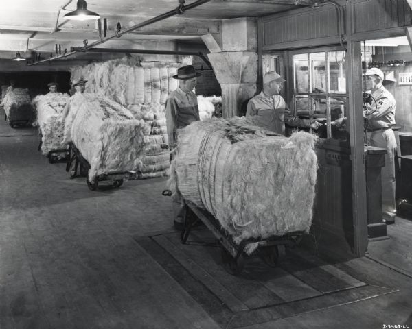 Factory workers weigh a large bale of Sisal fiber (fibre) at an International Harvester twine mill. Original caption reads: "Weighing the bales of Sisal fiber as they are unloaded from the cars and put into the warehouse for storage until ready to use."