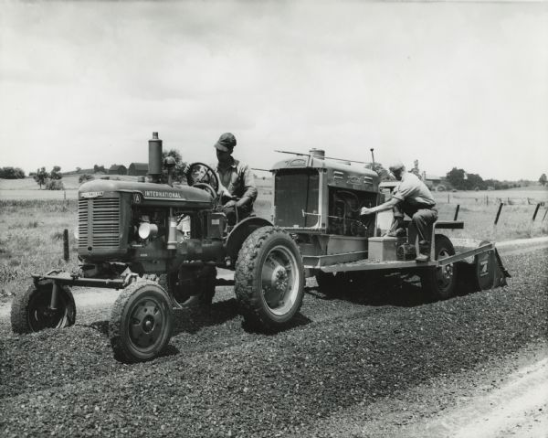 men spread asphalt on a road using an International A tractor and Seaman asphalt mixer. Original caption reads: "An International A being used to haul a Seaman asphalt mixer. Ordinarily the unit can be operated by the tractor operator. However, in this case the unit was new and needed minor adjustments during the operation. Spreading asphalt on 1-8/10 miles required 10 hours."