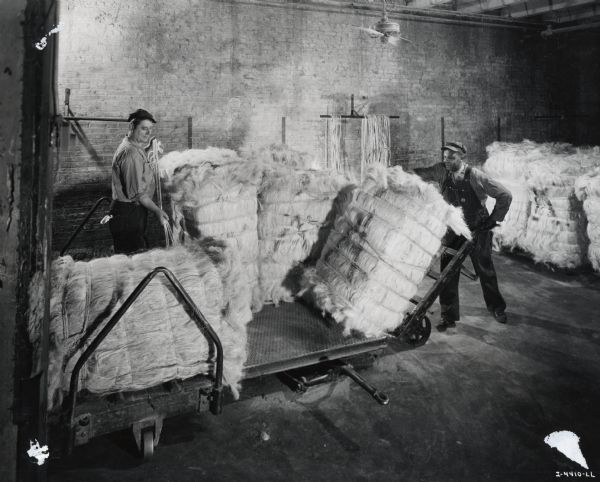 Two factory workers break bales of sisal in a warehouse. Caption on photograph reads: "Breaking the bales before putting them through the blending machines."
