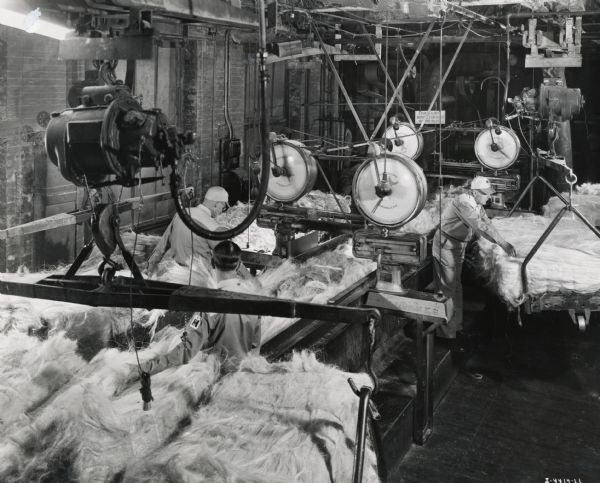Men use machines to blend fiber for binder twine at an International Harvester twine mill (most likely the McCormick Twine Mill). Original caption reads: "'Blending the Fiber.' Each of the three men shown at work in this illustration has a different kind of fiber on his truck. Each must feed fiber in proper relation to the other two in order to maintain the correct blend. Scales pictured govern the amount of fiber fed onto moving conveyor of this machine, shown as the first breaker or combing machine. Each scale has two pointers which indicate if the fiber is being fed correctly."