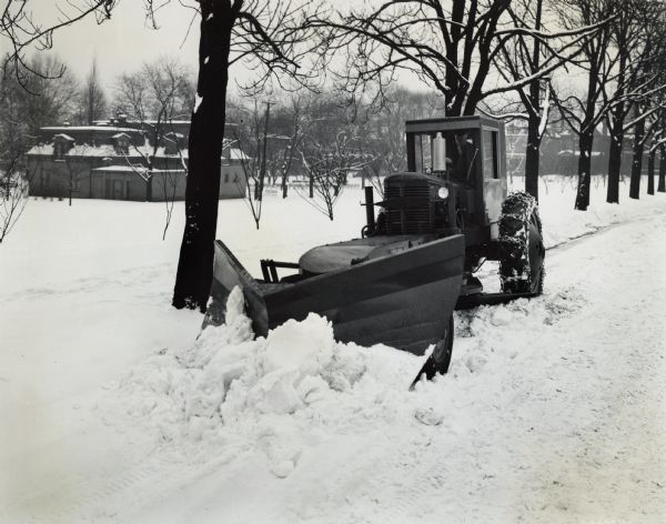 International (or Farmall?) tractor with Trojan Vee-plow removing snow in Pittsburgh. Original caption reads: "In a recent big snow storm which snarled Pittsburgh traffic, a Trojan one-man speed patrol equipped with Trojan Vee-plow was put to work on Thomas Boulevard. It is in front of the Old Westinghouse estate, which was willed to the city of Pittsburgh by George Westinghouse and is known as Westinghouse Park. The former stables are in the background. The regular 10-foot maintaining blade is in down position supplementing the clearing action of the Vee-plow. The Trojan patrol, which is powered by International M tractor, is a fast-moving one-man maintainer, its speeds varying from two to sixteen miles per hour."

