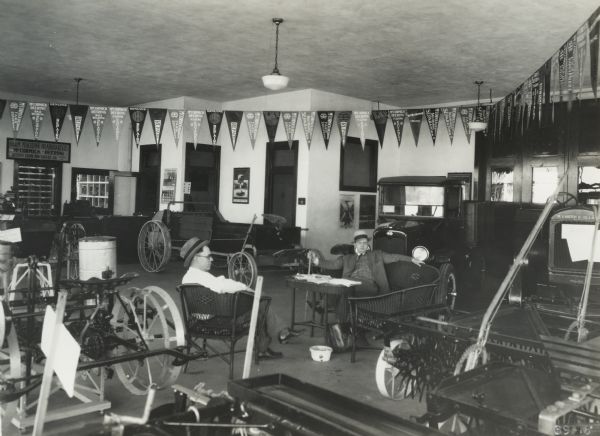 Two men sit on wicker furniture in the showroom of the Ottawa Implement Company, an International Harvester dealership. The showroom filled with tractors, cars, wagons, and farm machinery. Pennants cross the ceiling and advertising posters are mounted on the walls. A sign on the left wall reads: "Farm Machine Headquarters McCormick-Deering Ottawa Farm Machinery Co. Ottawa Ill." Both men are wearing hats, and the far one, facing the viewer is holding a cane.