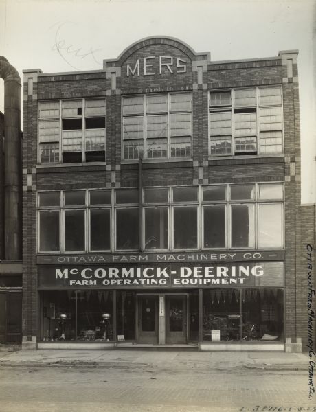 Exterior of the Ottawa Farm Machinery Company, an International Harvester dealership. Agricultural equipment and machinery are visible through the storefront windows on the ground level. The words "McCormick-Deering-Farm Operating Equipment" are painted on the glass.  On the top of the building is a decorative arch with the letters MERS.