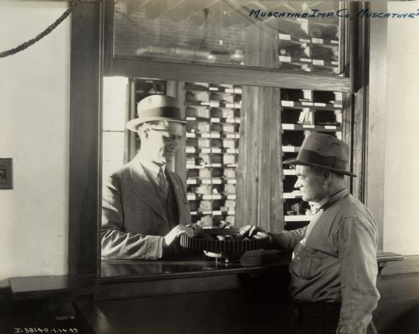 Service or parts counter at the Muscatine Implement Company, an International Harvester dealership. A man, possibly a customer, is standing at the counter with his hand on a large gear. Another man, likely the owner of the dealership, is standing on the other side of the counter. Parts bins are in the background.