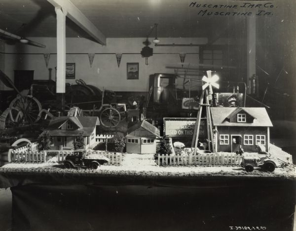 A tabletop display of a miniature farm, including a house, barn, garage, windmill, fences, plants, tractor, truck, animals, and two water pumps. A sign on the back of the table reads "McCormick-Deering Farm Machines Used on this Farm." In the background are tractors, trucks, and other farm equipment on display in the showroom of the Muscatine Implement Company, an International Harvester dealership.