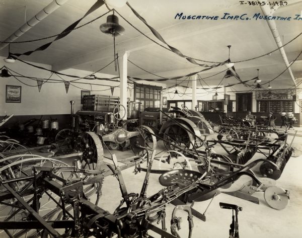 Tractors, plows, harrows and other farm equipment on display in the showroom of the Muscatine Implement Company, an International Harvester dealership. Banners and posters are displayed on the walls. There are two men sitting in wicker chairs in front of the service window near the back of the room.