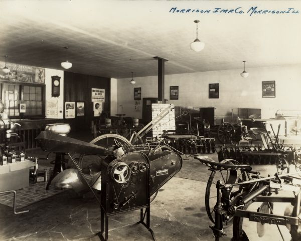 A corn sheller, plow and disc harrows on display in the showroom of the Morrison Implement Company, an International Harvester dealership. Posters and banners are on the walls and above the windows.