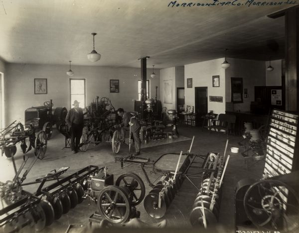 Two men in the showroom of the Morrison Implement Company, an International Harvester dealership. On display are tractors, plows, harrows, a stationary engine and other farm equipment. Also on display are advertising posters, a literature rack, and a table with toys. In the background, through an open door, is a restroom.
