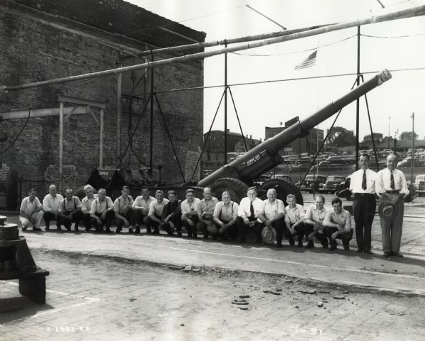 Milwaukee Works foremen and supervisors posing in front of a "high-speed, modernized gun carriage." At the right is Superintendent V.A. Guebard, and next to him is Menno Felber, second assistant superintendent. Two men are holding hats. A U.S. flag and a parking lot are behind the gun carriage.