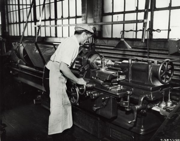 Factory worker at a machine in International Harvester's Milwaukee Works. Original caption reads: "High Speeding of 155-Millimeter Gun Carriage at Milwaukee Works. In this operation the ends of the rear axle of the 155-millimeter gun carriage are turned to the proper dimensions for the fitting into the rebuilt carriage capable of higher speeds. The turning is to reduce the size of the axle shaft for the wheels used on the higher speed carriage."