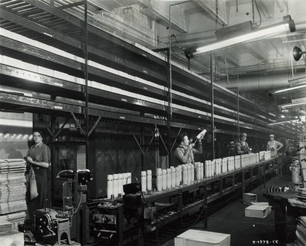 Factory workers producing artillery shells at International Harvester's Milwaukee Works. Original caption reads: "75-Millimeter Shell Manufacture. The shell conveyor line on which the shells are placed to dry after coming from the paint spray. Above and to the rear is a combination conveyor and storage unit for the finished shells, consisting of five conveyors at different levels. An automatic elevator carries the shell packages to the proper conveyor level and places them on the conveyor where they may move immediately to the shipping dock or remain in storage on the conveyor until such time as a sufficient stock has been accumulated for shipping."