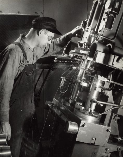 Factory worker wearing saftey goggles and welding a shell casing at International Harvester's Milwaukee Works. Original caption reads: "75-Millimeter Shell Manufacture. The base cover of the shell is welded in place on a spot welding machine to reinforce the shell against the force of the explosion."