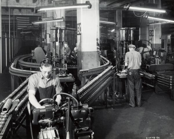 Factory workers produce artillery shells at International Harvester's Milwaukee Works. Original caption reads: "75-Millimeter Shell Manufacture. The conveyor equipment designed and installed by the Harvester Company in its shell machining department at Milwaukee Works to facilitate the rapid movement of shells from the machines to the first government inspection position. A Sundstrand machine for putting four notches in the end of the shell is shown in the foreground."