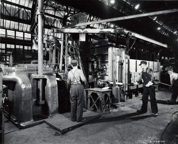 Factory workers producing artillery shells at International Harvester's Milwaukee Works. Original caption reads: "75-Millimeter Shell Manufacture. First operation in the production of 75-millimeter shells at the International Harvester Company's Milwaukee Works. Here steel rods are heated in a furnace and then chipped of loose particles of steel, after which the operator on the left places the bar in an upsetter machine. The bar is held in five different positions in the upsetter, after which the rough forging is completed. The forging is then lifted by the operator in the upper left of the photograph and placed on a conveyor to carry it to the next operation."