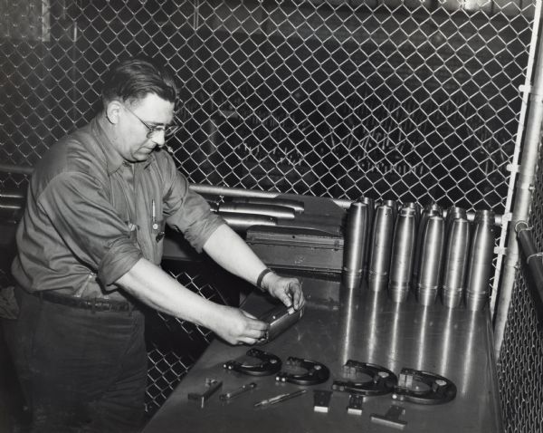 Factory inspector examining artillery shells at International Harvester's Milwaukee Works. Original caption reads: "Inspector Thomas Pedersen checks 75 mm shell casings manufactured there for correct size. The shells are complete except for welding the cushion plate on the bottom and painting. These shells are painted yellow before shipment to a government arsenal and red after they have been loaded."