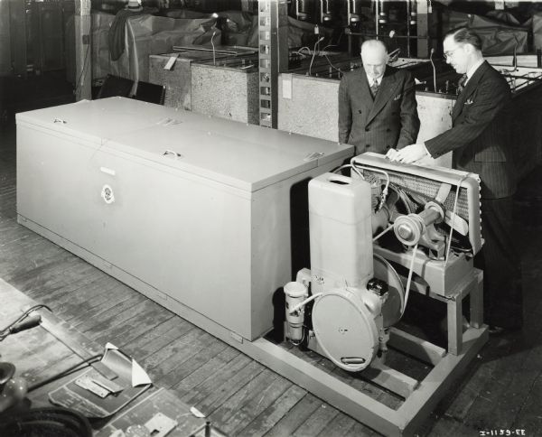 West Pullman Works managers standing with a refrigerator produced for the U.S. Marines. Original caption reads: "International, self-contained, engine-driven, portable. Sold to U.S. Marines and delivered to the Philadelphia Navy Yard March 19, 1941. No. 12 cooler cabinet with International 1.5 HP condensing Unit operated by La 1.5-2.5 HP engine. Has approximate 50 cubic foot capacity and will hold approximately a ton of meat around 36 degrees F. Mr. Watling (left) superintendent of West Pullman Works and Mr. Johnson (assistant superintendent, right)-standing behind the unit."