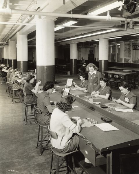 Factory workers inspecting aircraft torpedo parts at an International Harvester factory. Original caption reads: "Women are employed by the company to inspect the many hundreds of small parts used in the torpedo. They work with small precision gauges and micrometers. All are new employees trained for this special work. Prior to becoming torpedo inspectors they were tool crib attendants, dress designers, writers, etc."