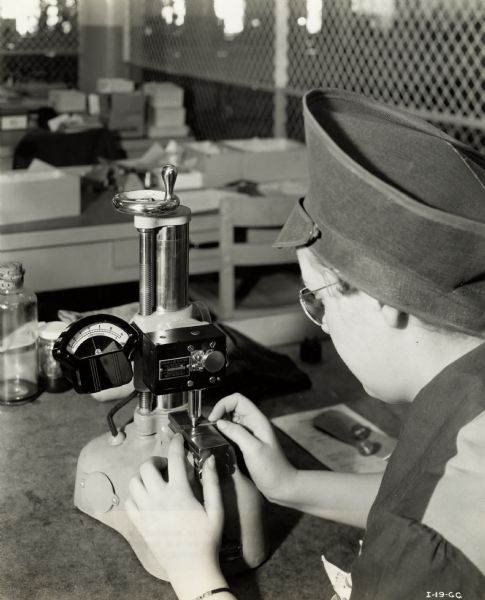 Female worker inspecting a ball bearing for an aircraft torpedo at an International Harvester factory. Original caption reads: "The precision instrument used in measuring ball bearings for a torpedo part. The inspector pushes a tiny bearing under the indicator and the gauge on the left measures to a fineness of .000025 or 25 millionths of an inch."